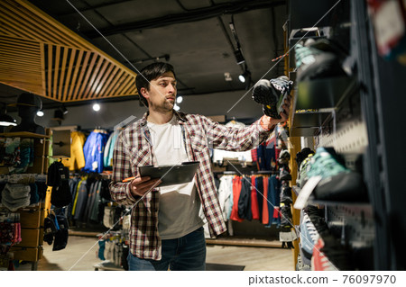 Sporting goods store owner with clipboard checking inventory. Tourist store manager working near showcase with hiking boots makes check list of orders. Salesman exhibit display of walking boots Sporting goods store owner with clipboard checking inventory. Tourist store manager working near showcase with hiking boots makes check list of orders. Salesman exhibit display of walking boots 76097970