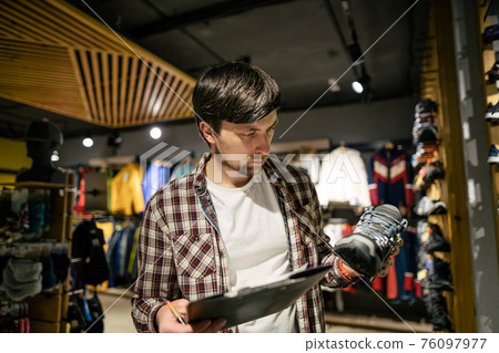 Sales assistant sports travel store makes inventory hiking boots. Travel goods store theme. Clothes and footwear for sports and travel. Store manager near shelves with hiking boots makes check list 76097977