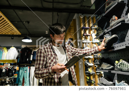 Business owner in sports store works with showcase trekking and mountaineering boots for sale, holding clipboard in his hands, checks availability of goods and collects to process online orders 76097981