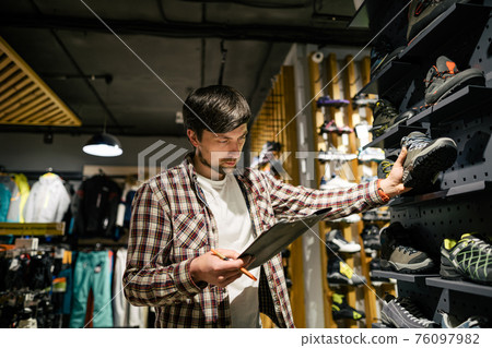 Sporting goods store owner with clipboard checking inventory. Tourist store manager working near showcase with hiking boots makes check list of orders. Salesman exhibit display of walking boots 76097982