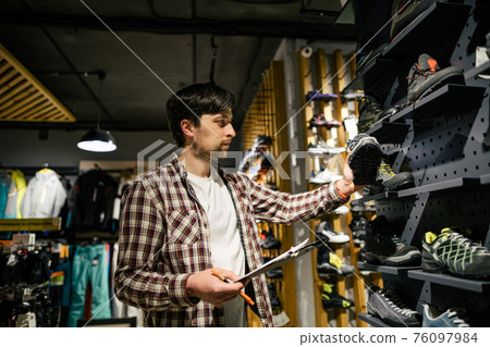 Business owner in sports store works with showcase trekking and mountaineering boots for sale, holding clipboard in his hands, checks availability of goods and collects to process online orders 76097984