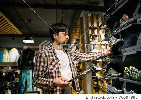 Sporting goods store owner with clipboard checking inventory. Tourist store manager working near showcase with hiking boots makes check list of orders. Salesman exhibit display of walking boots 76097985