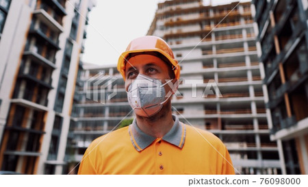 Topic is health protection in production and construction. Caucasian male worker in orange uniform and protective respirator kn 95 puts on construction orange hard hat and poses at construction site Topic is health protection in production and construction. Caucasian male worker in orange uniform and protective respirator kn 95 puts on construction orange hard hat and poses at construction site 76098000