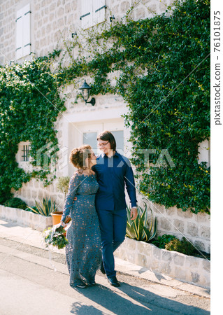 The bride and groom are embracing along the road near an old building with a white door, the wall is entwined with a green liana  76101875
