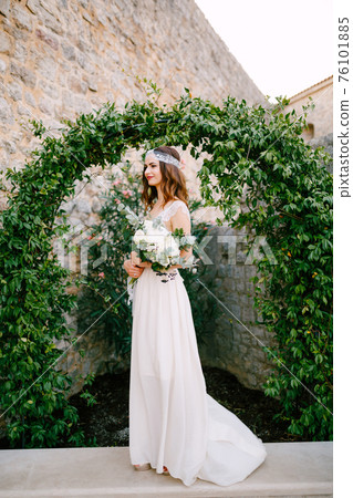 A bride stands at a graceful arch entwined with wild grapes in the old town of Budva and holds a bouquet in her hands  76101885