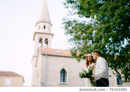 The bride and groom walk through the old town of Budva, the groom gently hugs the bride and holds a bouquet in his hand  76101980
