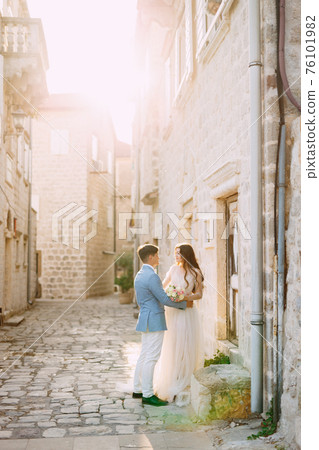 The bride and groom hug on a beautiful old street of Perast near a white stone wall The bride and groom hug on a beautiful old street of Perast near a white stone wall 76101982