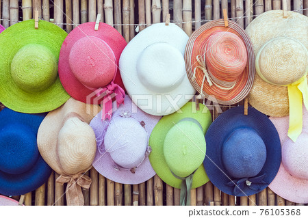 A variety of fashionable and colorful hats hanging on the bamboo backdrop of curbside shop.Women's designer hats from the sun of different colors. Design of women's beach hats. Beach hats for summer. 76105368