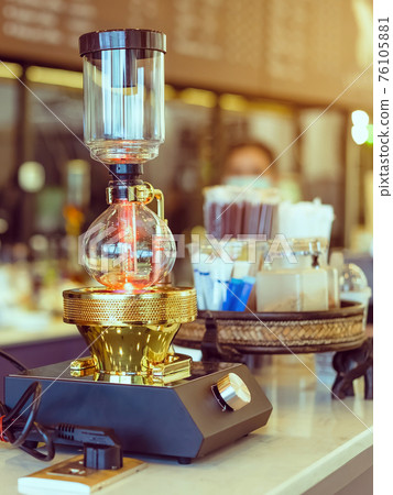 Close up to barista make coffee with Vacuum Pot Syphon Brewer. Syphon (Siphon) coffee maker brews drink using two chambers where vapor pressure and vacuum produce coffee in cafe. Selective focus. Close up to barista make coffee with Vacuum Pot Syphon Brewer. Syphon (Siphon) coffee maker brews drink using two chambers where vapor pressure and vacuum produce coffee in cafe. Selective focus. 76105881