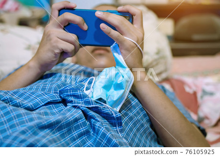 A surgical mask to prevent  infection of Coronavirus(C0VID-19) hung on hand of young man while lying on the bed to play game with mobile phones. New normal lifestyle. Selective focus on surgical mask. 76105925