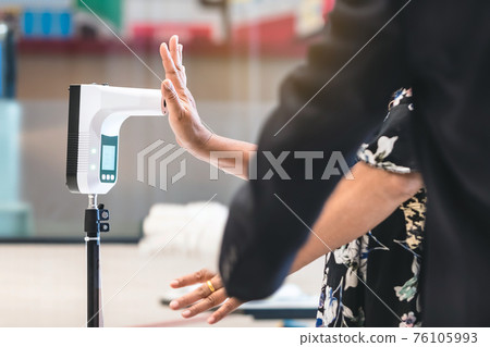 Customer with protective mask raised hand at digital thermometer with temperature scanning machine for fever before entering area. Screening to prevent infected people from entering restricted areas. Customer with protective mask raised hand at digital thermometer with temperature scanning machine for fever before entering area. Screening to prevent infected people from entering restricted areas. 76105993