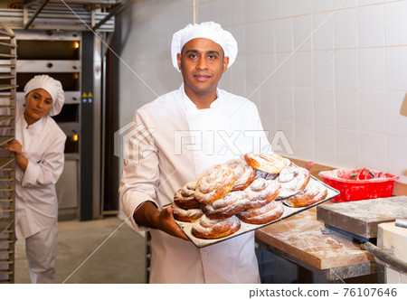 Male baker holding tray of hot sweet pastry in the kitchen of the bakery 76107646