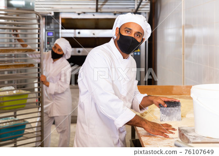 Bakery worker in protective mask cuts raw dough with knife to prepare for baking Bakery worker in protective mask cuts raw dough with knife to prepare for baking 76107649