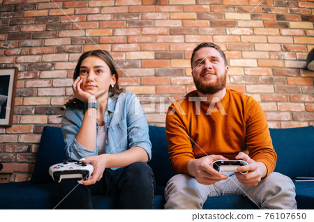 Front view of happy cheerful young couple holding controllers and playing video games on console sitting together on couch Front view of happy cheerful young couple holding controllers and playing video games on console sitting together on couch 76107650