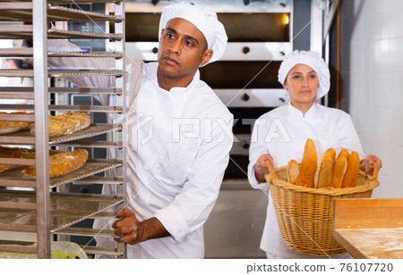 Focused baker pushing rack trolley with baked baguettes 76107720