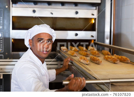Hispanic baker taking baked baguettes out of oven in bakeshop 76107820