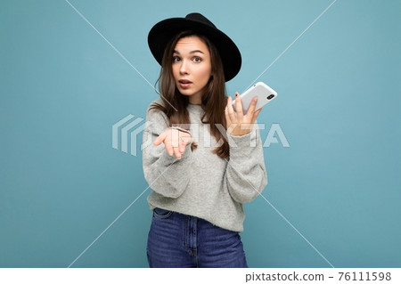 Beautiful young asking dissatisfied brunette woman wearing black hat and grey sweater holding 76111598