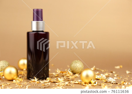 Unbranded dark brown spray bottle, gold Christmas balls and pieces of gold paper on golden background. Cosmetic packaging mockup with copy space, front view. Body mist. 76111636