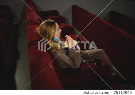 young woman in medical mask eating popcorn sitting in the cinema 76113949