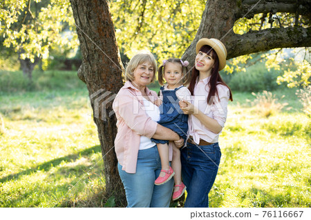Outdoor bright portrait of happy cheerful multi generation family, smiling grandmother, young mother and little cute 3 years old girl, posing to camera in the garden, standing under big tree 76116667