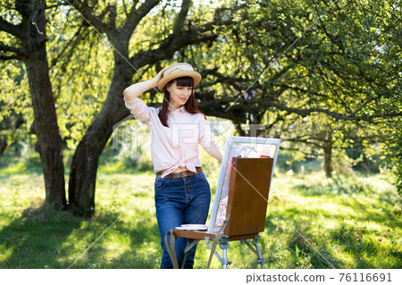Young Caucasian woman artist, wearing jeans, light shirt and straw hat, painting a picture on an easel in sunny summer park Young Caucasian woman artist, wearing jeans, light shirt and straw hat, painting a picture on an easel in sunny summer park 76116691