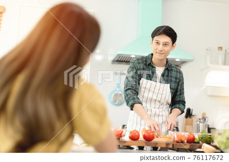 Husband slicing vegetables to prepare breakfast. With a young wife supporting him In a romantic atmosphere 76117672