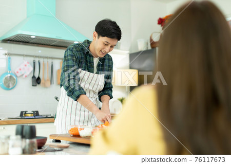 Husband slicing vegetables to prepare breakfast. With a young wife supporting him In a romantic atmosphere Husband slicing vegetables to prepare breakfast. With a young wife supporting him In a romantic atmosphere 76117673
