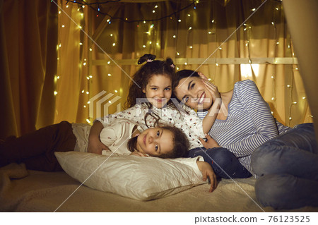 Affectionate mother and two little daughter relaxing in tent in home living room 76123525