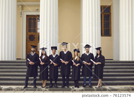 Group of smiling graduates standing with diplomas over university building background Group of smiling graduates standing with diplomas over university building background 76123574