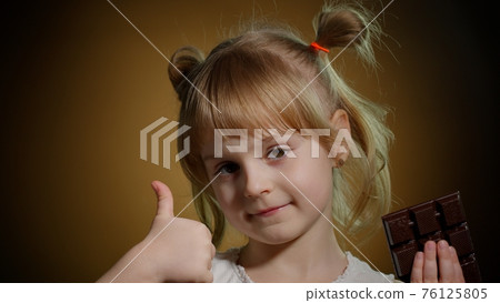Little child girl kid eating chocolate bar dessert, showing thumbs up isolated on dark background Little child girl kid eating chocolate bar dessert, showing thumbs up isolated on dark background 76125805