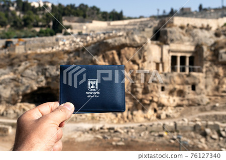 Repatriant's hand holds a temporary Israeli passport against the tomb of the priest Zechariah on the ancient jewish old cemetery on Mount of Olives in Jerusalem, Israel 76127340
