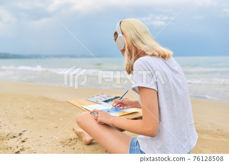 Young woman artist painting with watercolors sitting on the seashore Young woman artist painting with watercolors sitting on the seashore 76128508