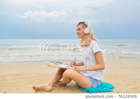 Young woman teenager in headphones drawing sketch with watercolors sitting on sea beach 76128509