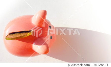 Top view over pink piggy bank with slot and inserted coin, on white background, savings and investment concept, copy space, space for text 76128591
