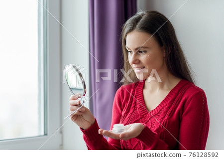 Young smiling woman with face moisturizer near eyes, female with mirror Young smiling woman with face moisturizer near eyes, female with mirror 76128592