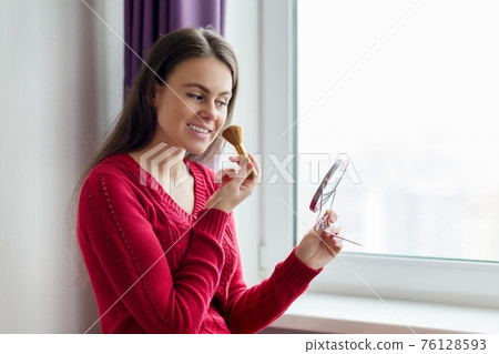 Young girl does makeup, stands near window with mirror and make-up brush Young girl does makeup, stands near window with mirror and make-up brush 76128593