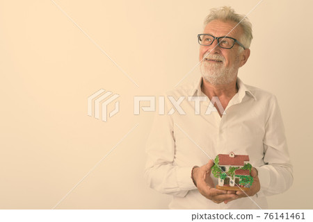 Studio shot of happy senior bearded man smiling while thinking and holding house figurine with 76141461
