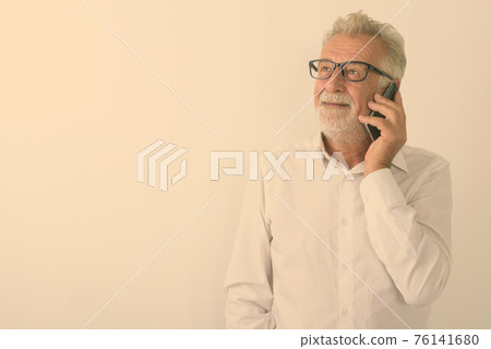 Studio shot of happy senior bearded man smiling and thinking while talking on mobile phone with 76141680