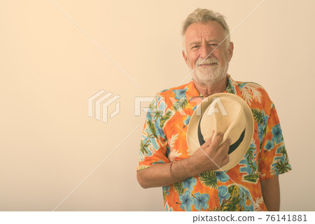 Studio shot of handsome senior bearded tourist man holding hat on his chest against white background 76141881