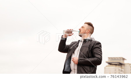 A man in a leather jacket drinks water from a bottle against the sky 76143962