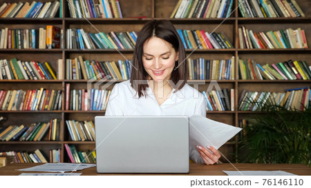 Young woman works with papers by laptop at table in library Young woman works with papers by laptop at table in library 76146110