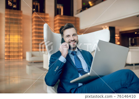 Man in a suit sitting in the arm-chair and working on a laptop Man in a suit sitting in the arm-chair and working on a laptop 76146410