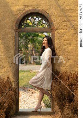 Happy young beautiful woman in soft white dress leaning on the door frame of her rustic garden green house on a bright sunny day 76157071