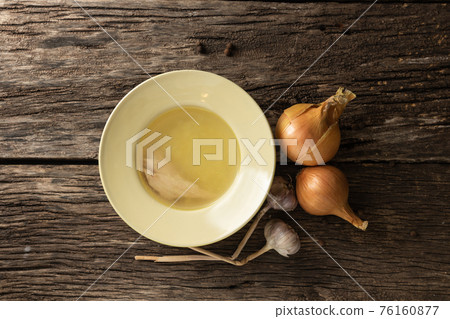 Bowl with clear rich broth. White crockery on an aged wooden table. Vegetables - onions, garlic, spices Bowl with clear rich broth. White crockery on an aged wooden table. Vegetables - onions, garlic, spices 76160877