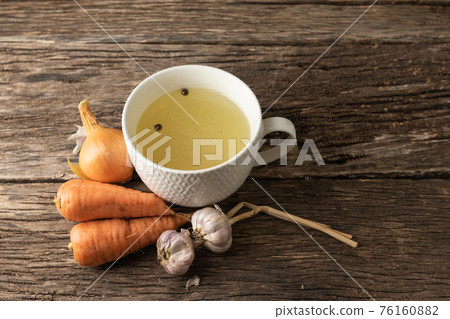 Bowl with clear rich broth. White crockery on an aged wooden table. Vegetables - carrots, onions, garlic, spices Bowl with clear rich broth. White crockery on an aged wooden table. Vegetables - carrots, onions, garlic, spices 76160882