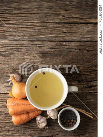 Bowl with clear rich broth. White crockery on an aged wooden table. Vegetables - carrots, onions, garlic, spices Bowl with clear rich broth. White crockery on an aged wooden table. Vegetables - carrots, onions, garlic, spices 76160884