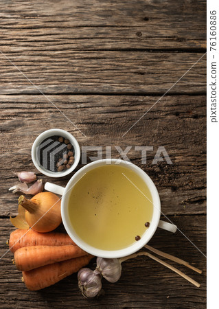 Bowl with clear rich broth. White crockery on an aged wooden table. Vegetables - carrots, onions, garlic, spices Bowl with clear rich broth. White crockery on an aged wooden table. Vegetables - carrots, onions, garlic, spices 76160886