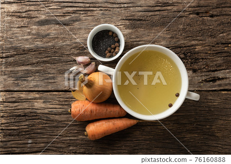 Bowl with clear rich broth. White crockery on an aged wooden table. Vegetables - carrots, onions, spices Bowl with clear rich broth. White crockery on an aged wooden table. Vegetables - carrots, onions, spices 76160888