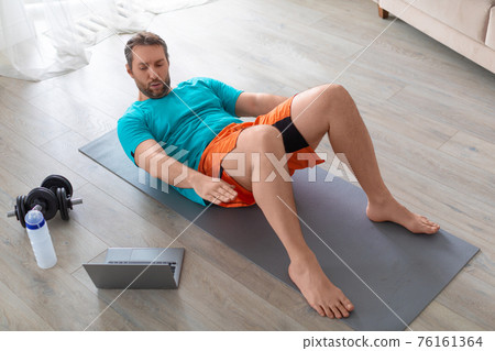 Middle aged man performs an abdominal exercise in front of a laptop monitor. He looks at the monitor and trains with an online guide. Middle aged man performs an abdominal exercise in front of a laptop monitor. He looks at the monitor and trains with an online guide. 76161364
