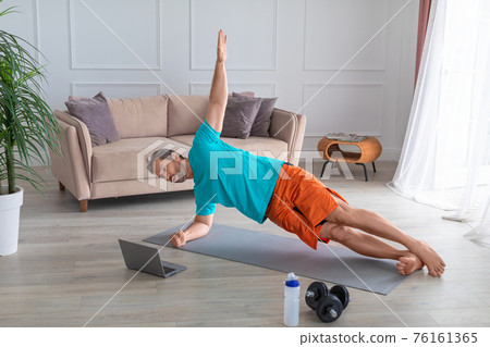 Workout at home. A middle-aged man performs an exercise in front of a laptop monitor - side plank. He trains with an online guide. Workout at home. A middle-aged man performs an exercise in front of a laptop monitor - side plank. He trains with an online guide. 76161365